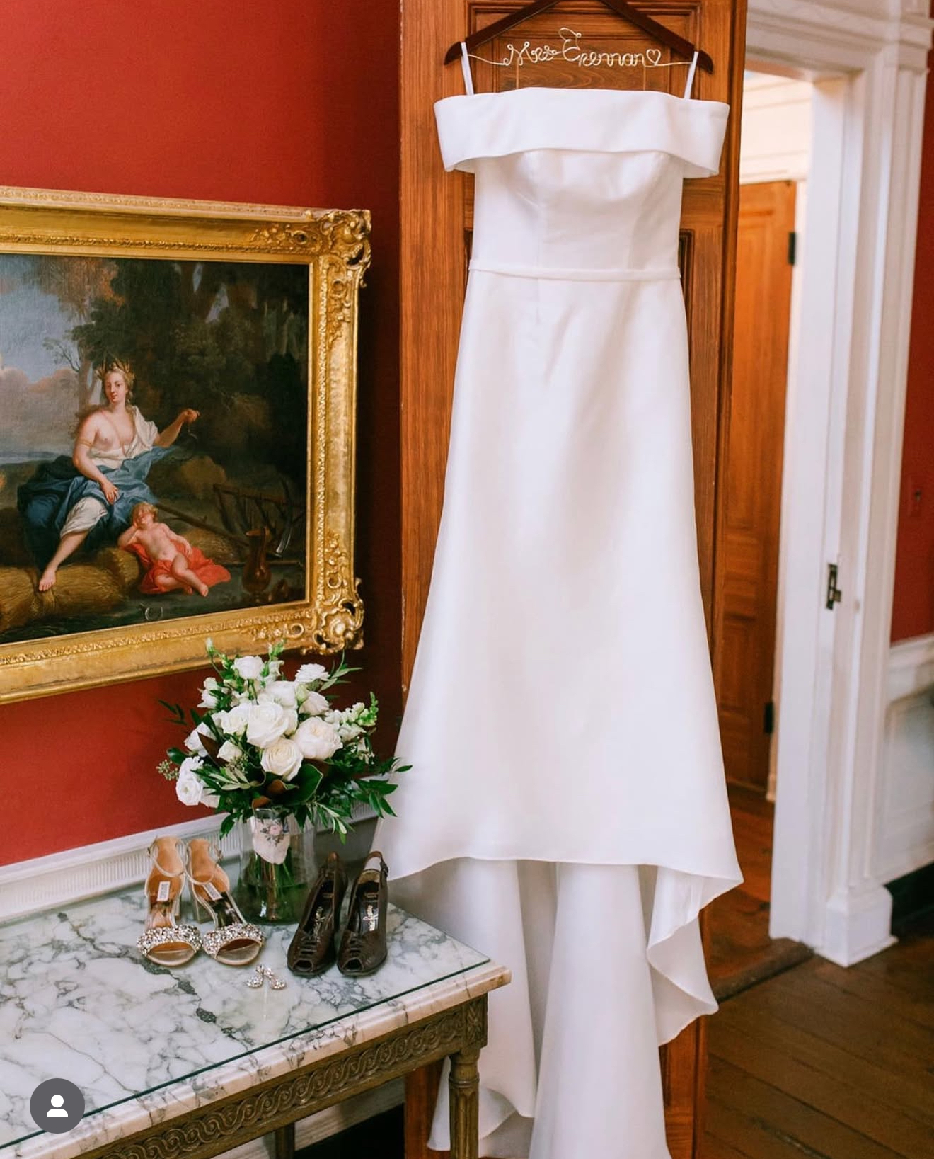 Off-the-shoulder wedding gown on a hanger beside a bridal bouquet and shoes